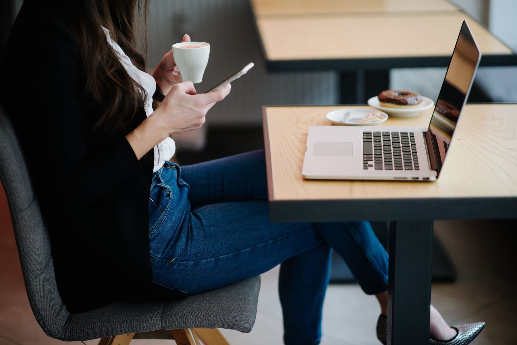Woman in a café holding coffee and typing on her smartphone beside a laptop