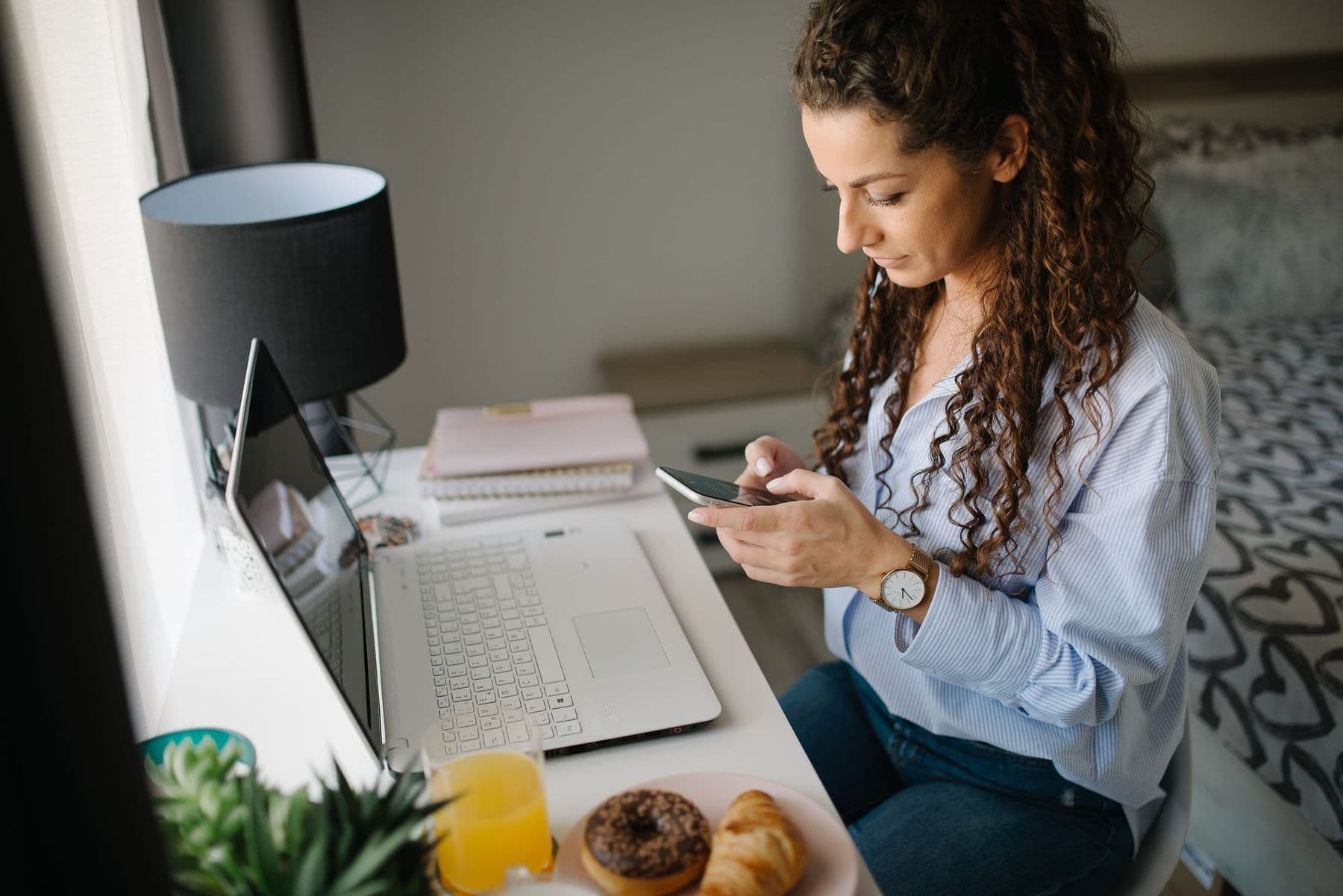 Woman at a desk reading messages on her smartphone next to a laptop