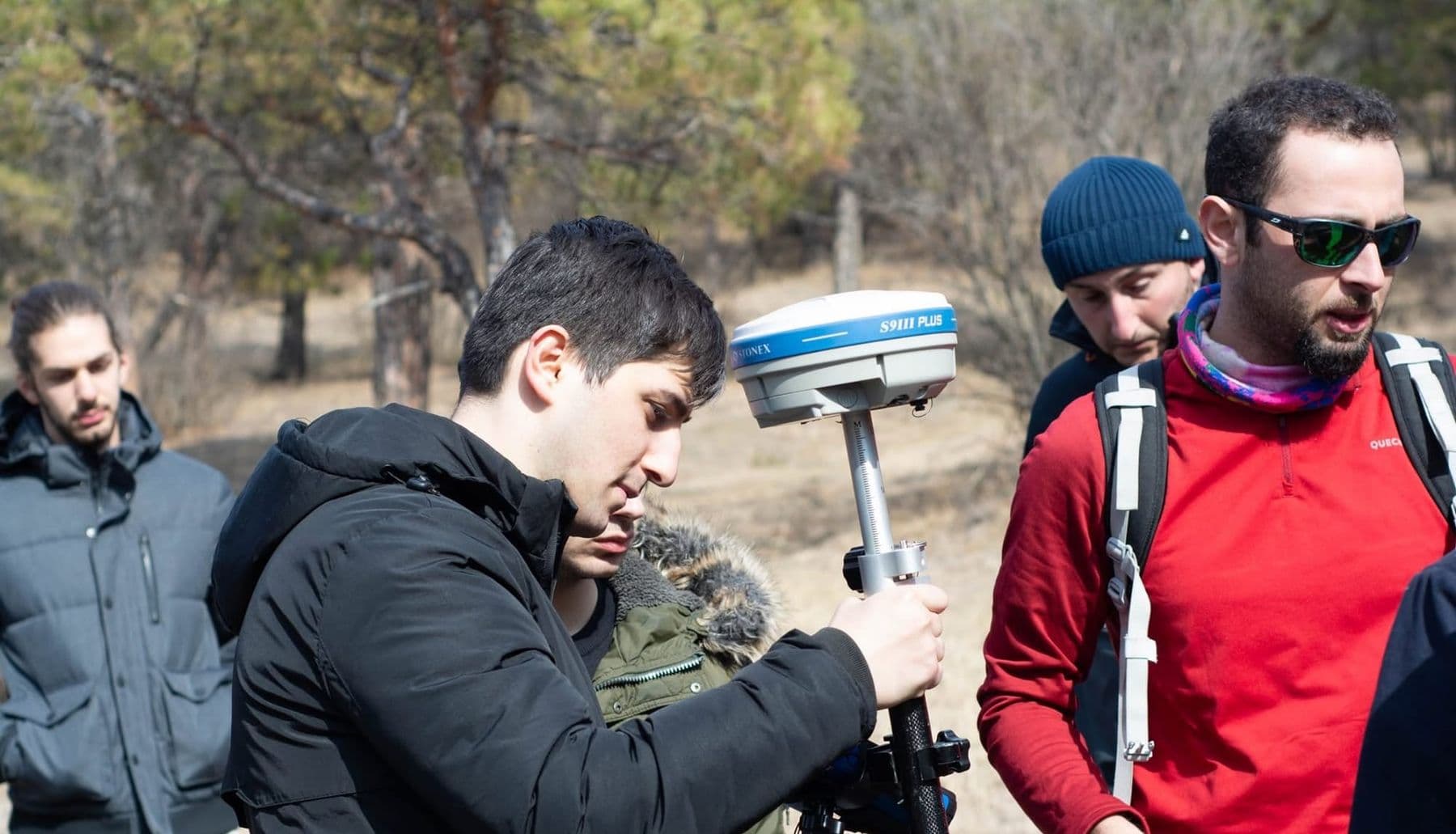 A young man checks a GNSS receiver pole outdoors while others watch