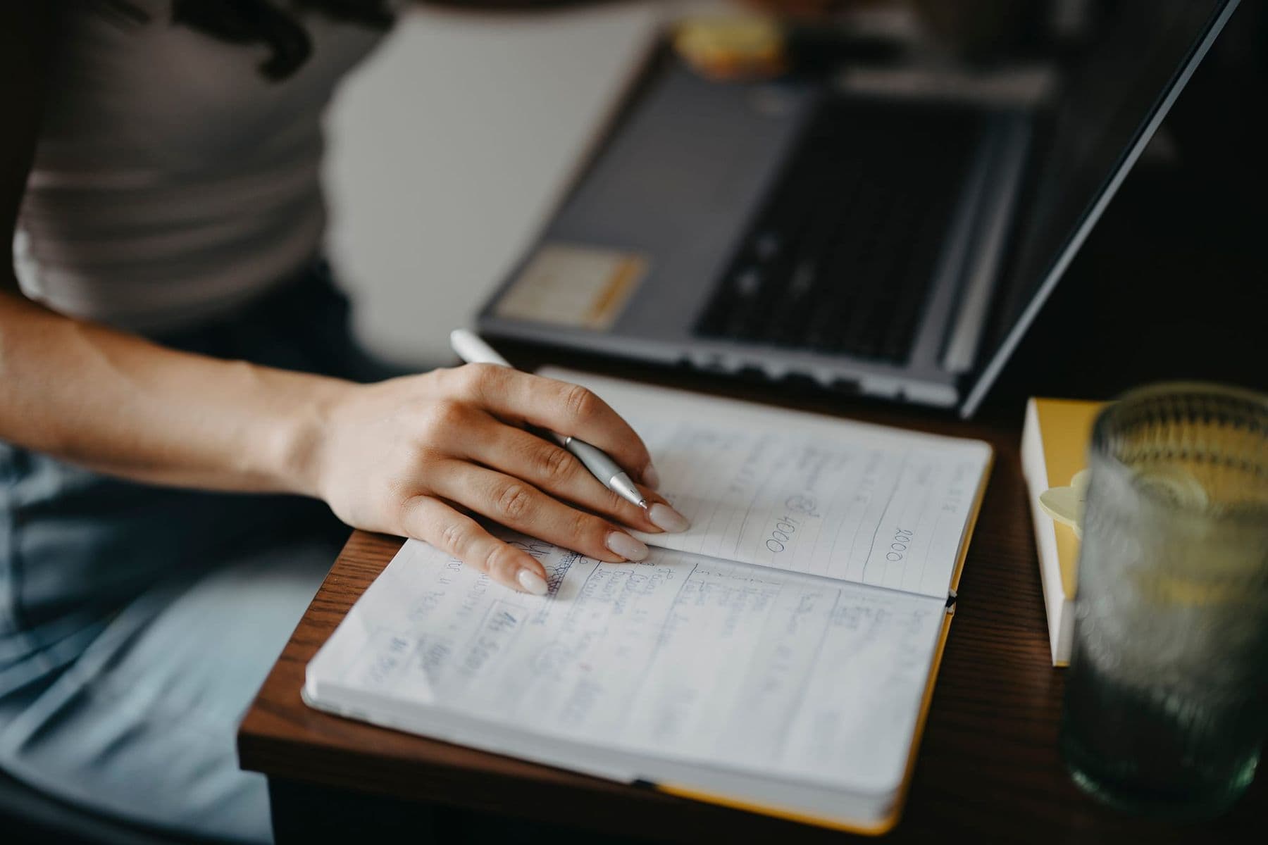 Teacher writing notes in a notebook next to a laptop while grading