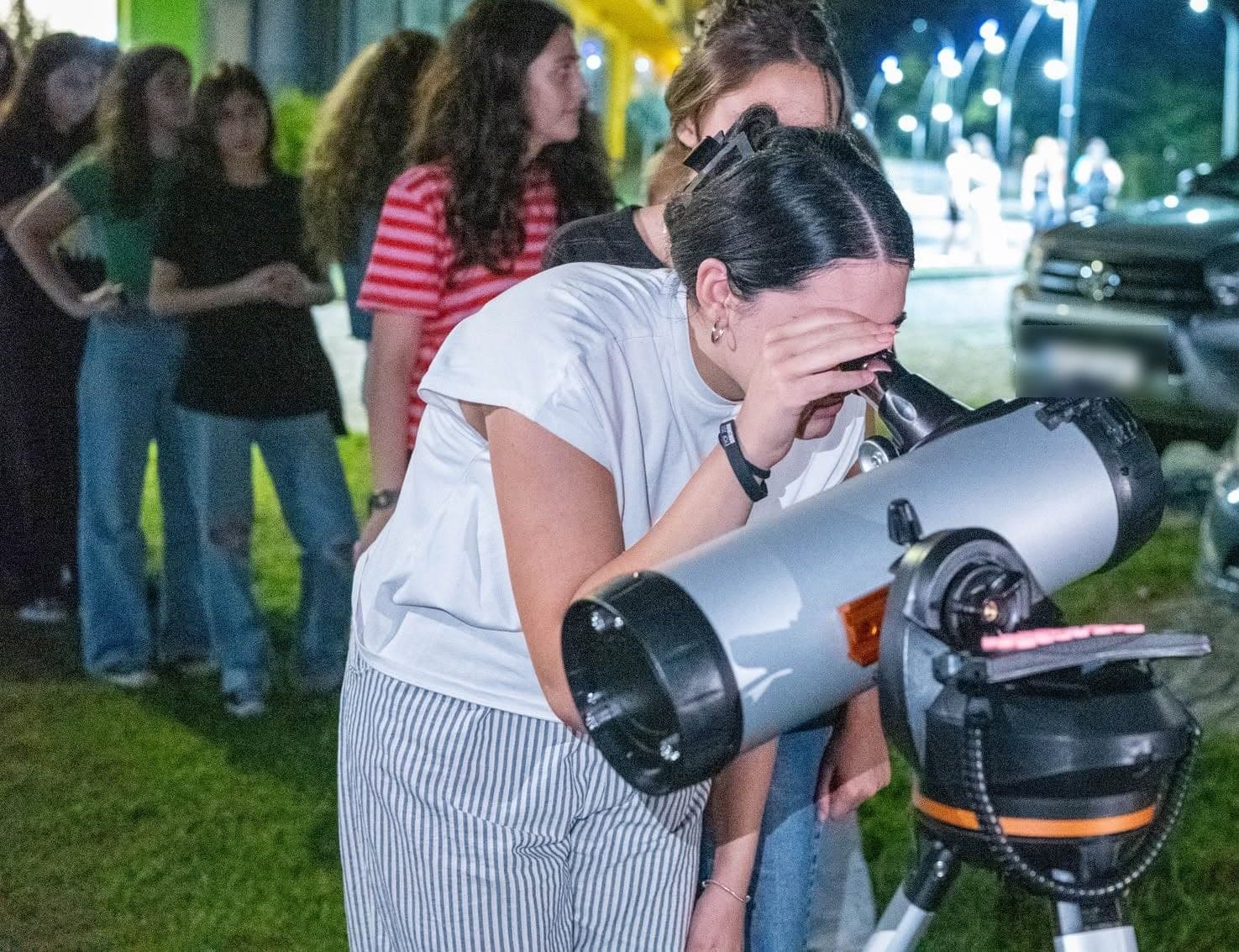 A teenage girl looks through a telescope at night while others wait behind her