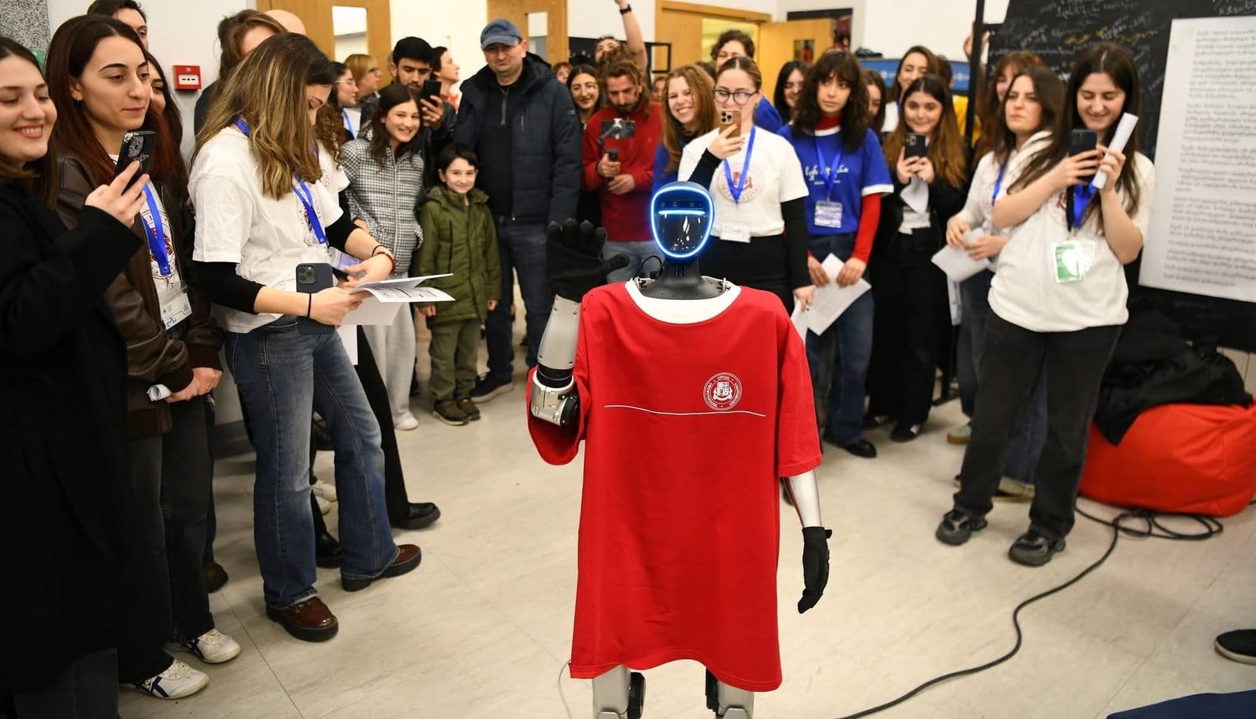 A humanoid robot in a red T-shirt faces young people filming it with phones