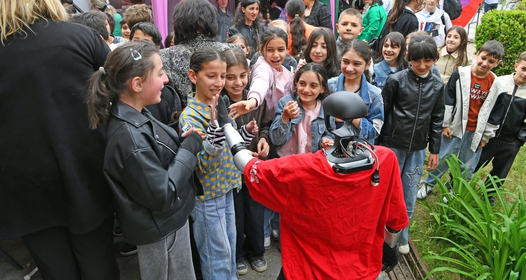 Excited children crowd around a humanoid robot in a red shirt outdoors